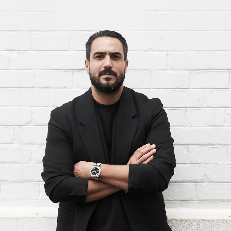 Portrait du Chef Pâtissier Jeffrey Cagnes en veste noire les bras croisés, adossé à un mur de briques blanches à Paris.