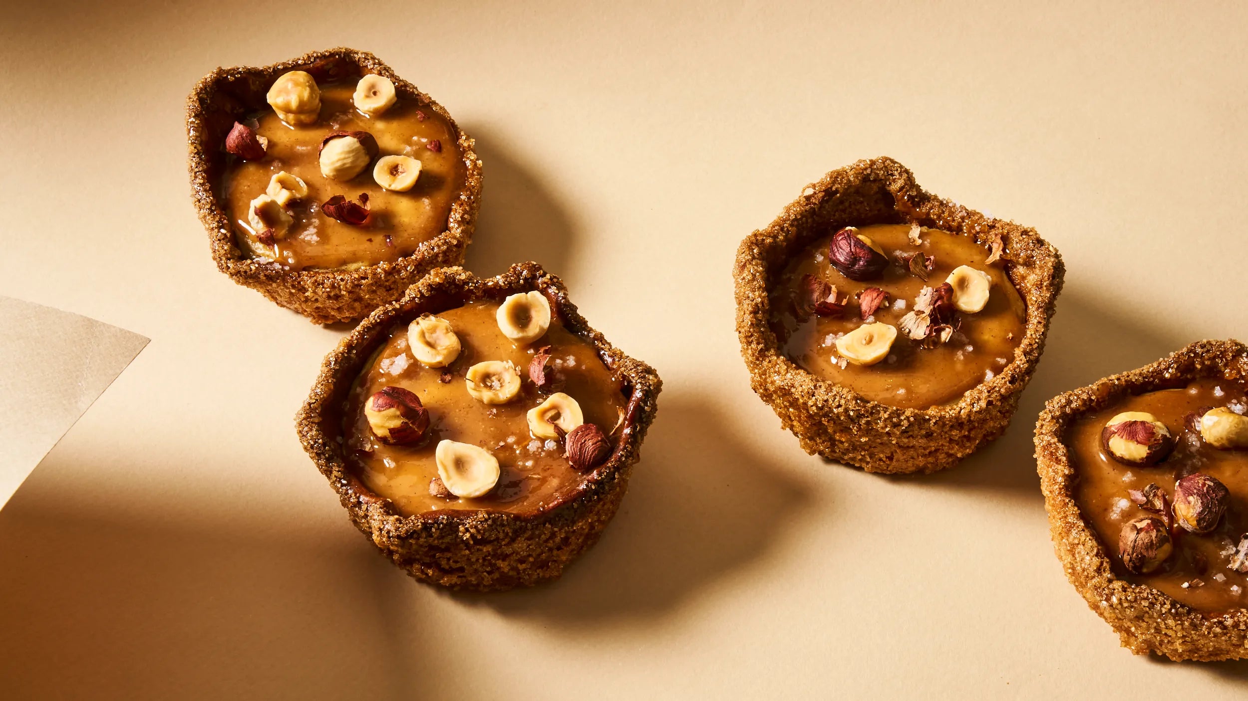 Vue de dessus de quatre flans pâtissiers noisette de Jeffrey Cagnes, disposés en diagonale sur un fond beige. Chaque tartelette présente une croûte rustique et est garnie de crème à la noisette caramélisée, parsemée de noisettes torréfiées entières et concassées.