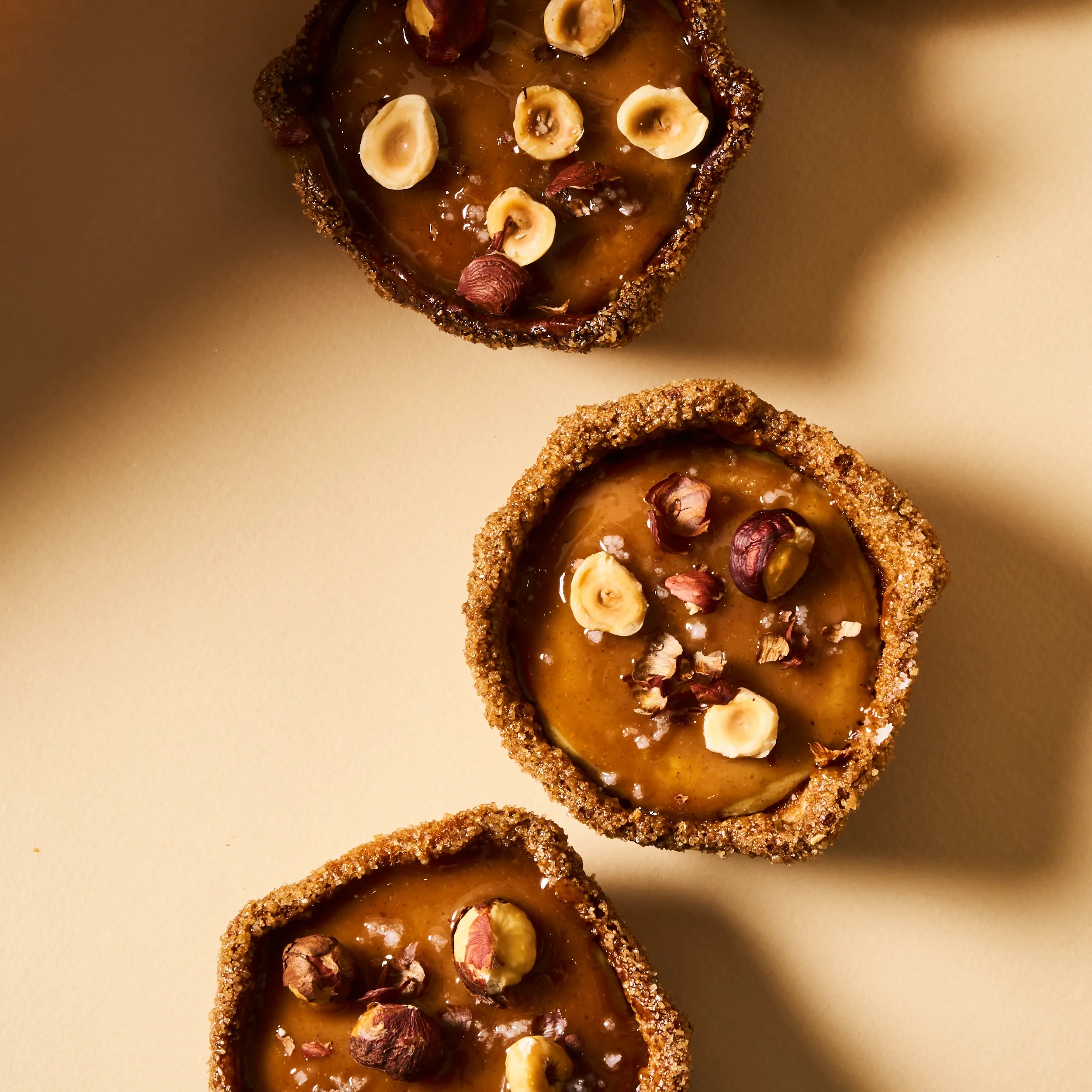 Trio de flans noisette individuels vus du dessus, parsemés de noisettes torréfiées entières et d'éclats de fleur de sel sur un praliné brillant.
