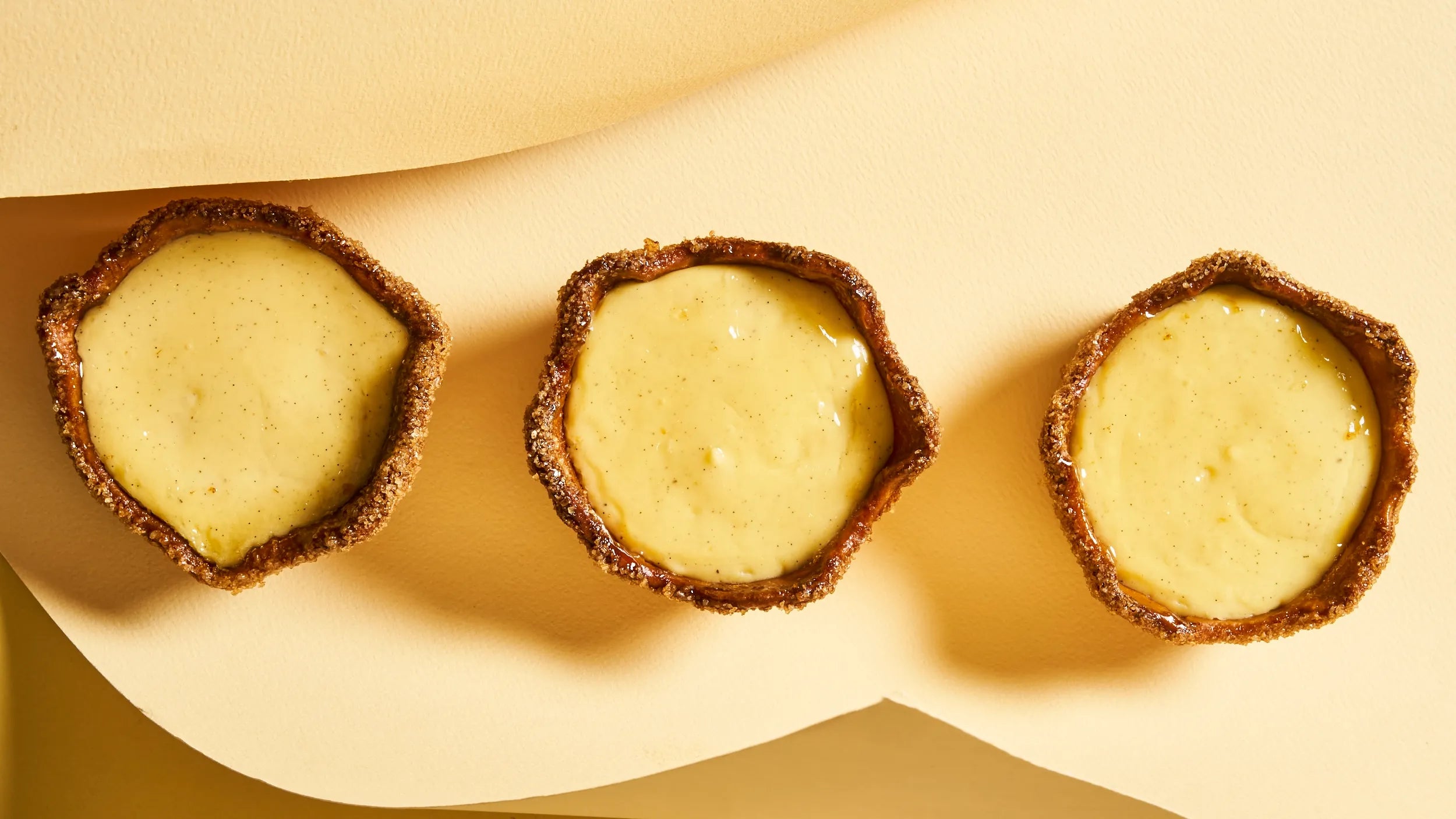 Trois flans individuels à la vanille de Madagascar vus de dessus, alignés sur un fond crème épuré, mettant en valeur leur appareil onctueux parsemé de grains de vanille.