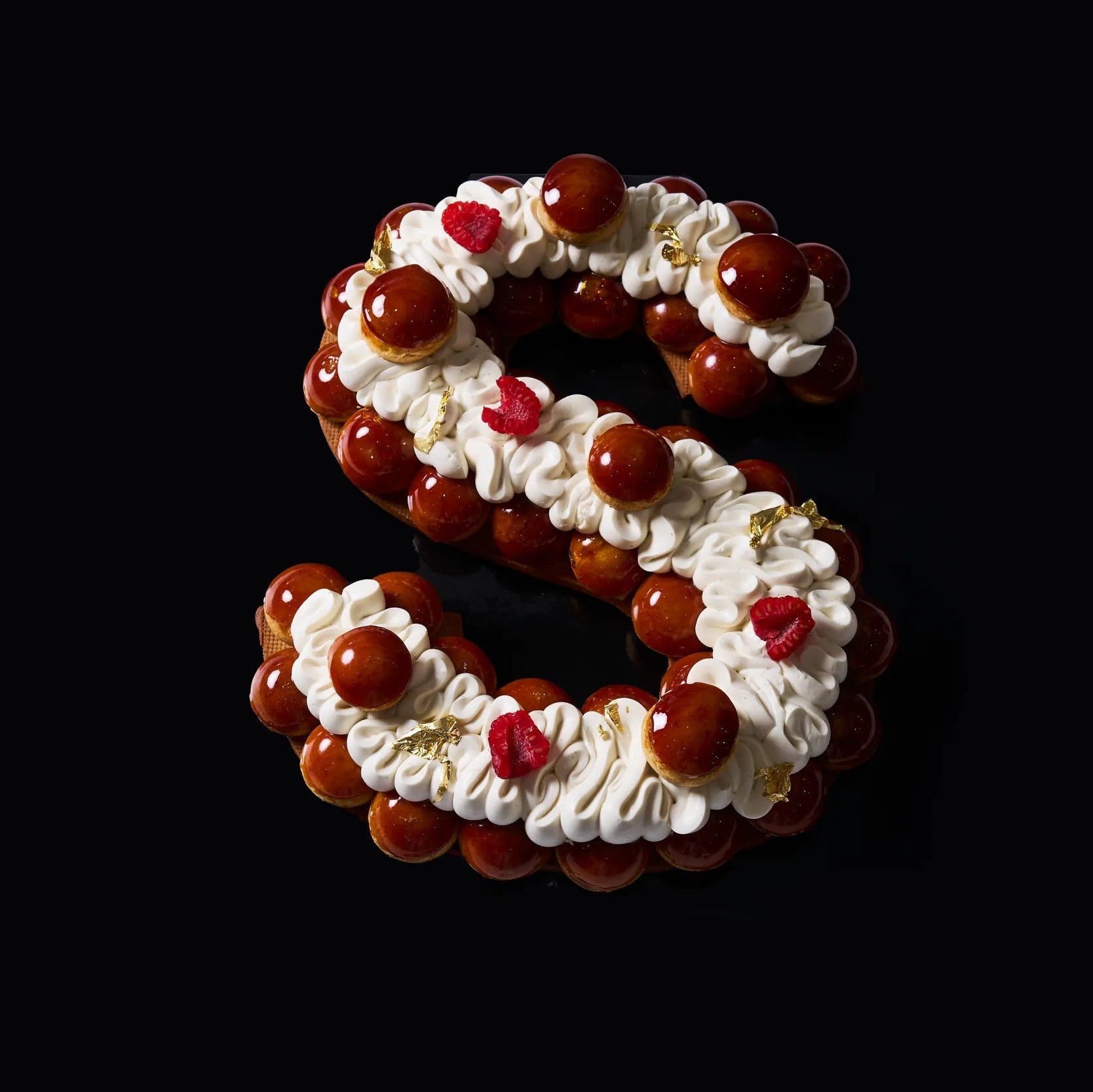 Letter cake d'exception en forme de lettre S façon Saint-Honoré par le Chef Jeffrey Cagnes, composé de choux caramélisés brillants, de crème pochée, de framboises fraîches et de feuilles d'or sur fond noir épuré.