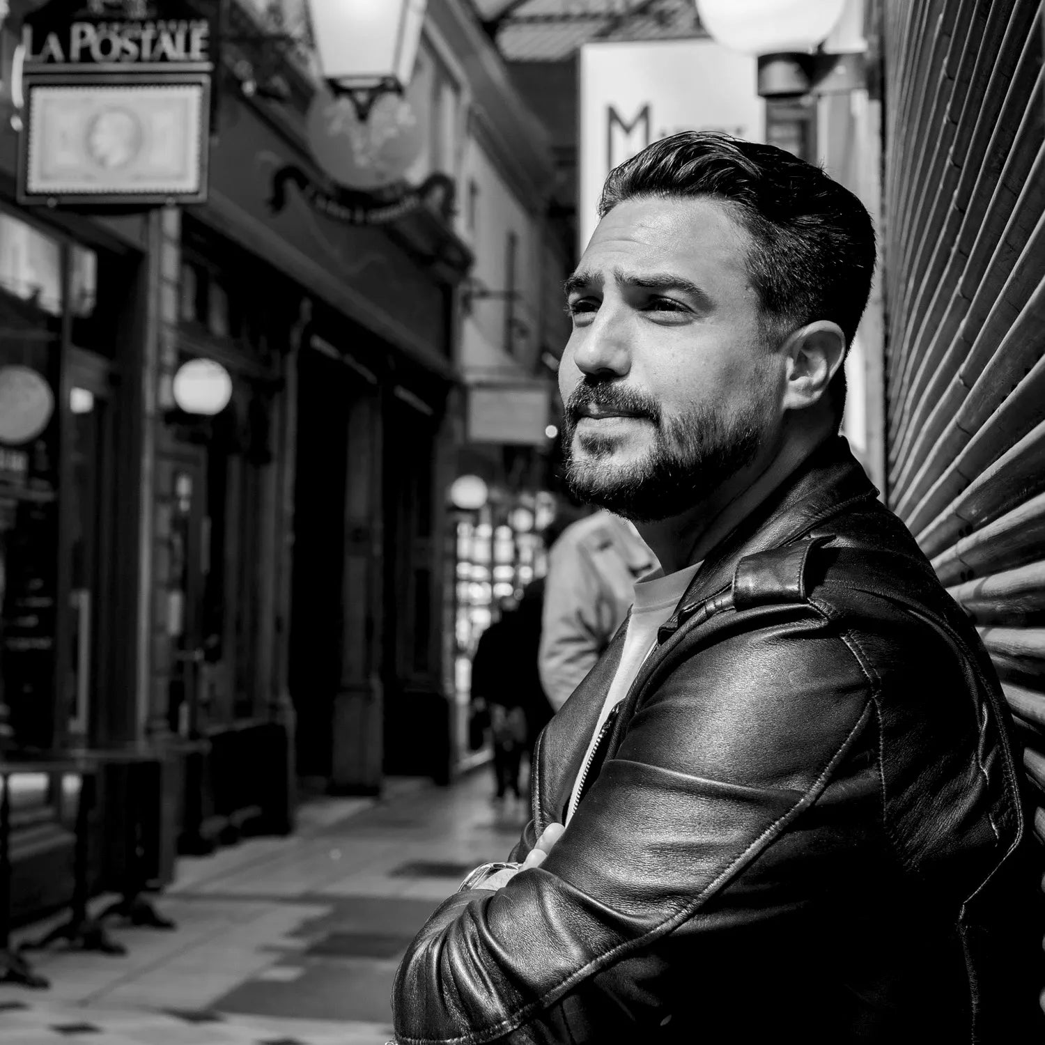 Portrait de Jeffrey Cagnes portant une veste en cuir noire, regardant au loin, debout dans le Passage des Panoramas à Paris, avec des enseignes historiques en arrière-plan.
