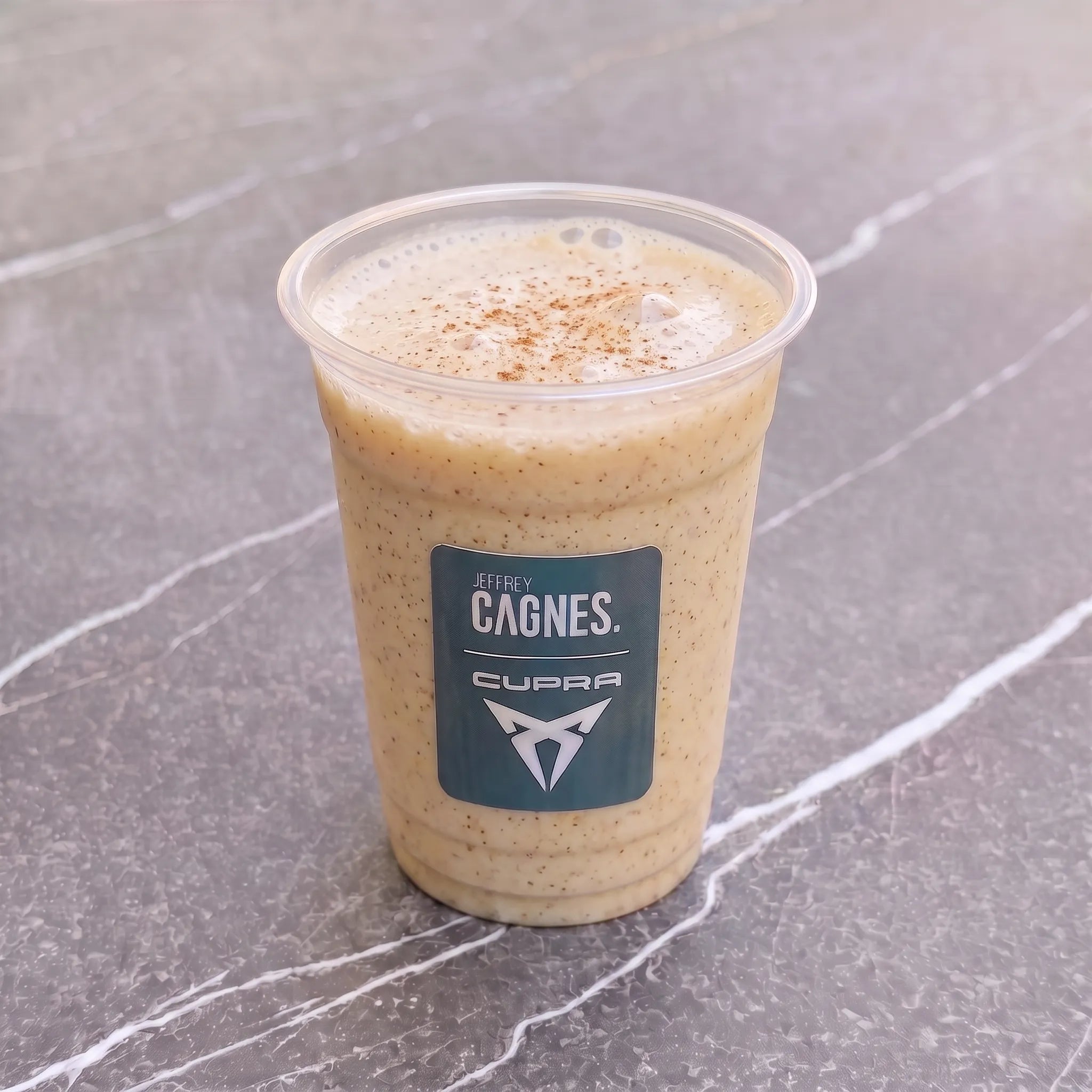 Gobelet transparent de smoothie Apple Cinnamon par Jeffrey Cagnes x Cupra, à la couleur ambrée et délicatement saupoudré de cannelle.