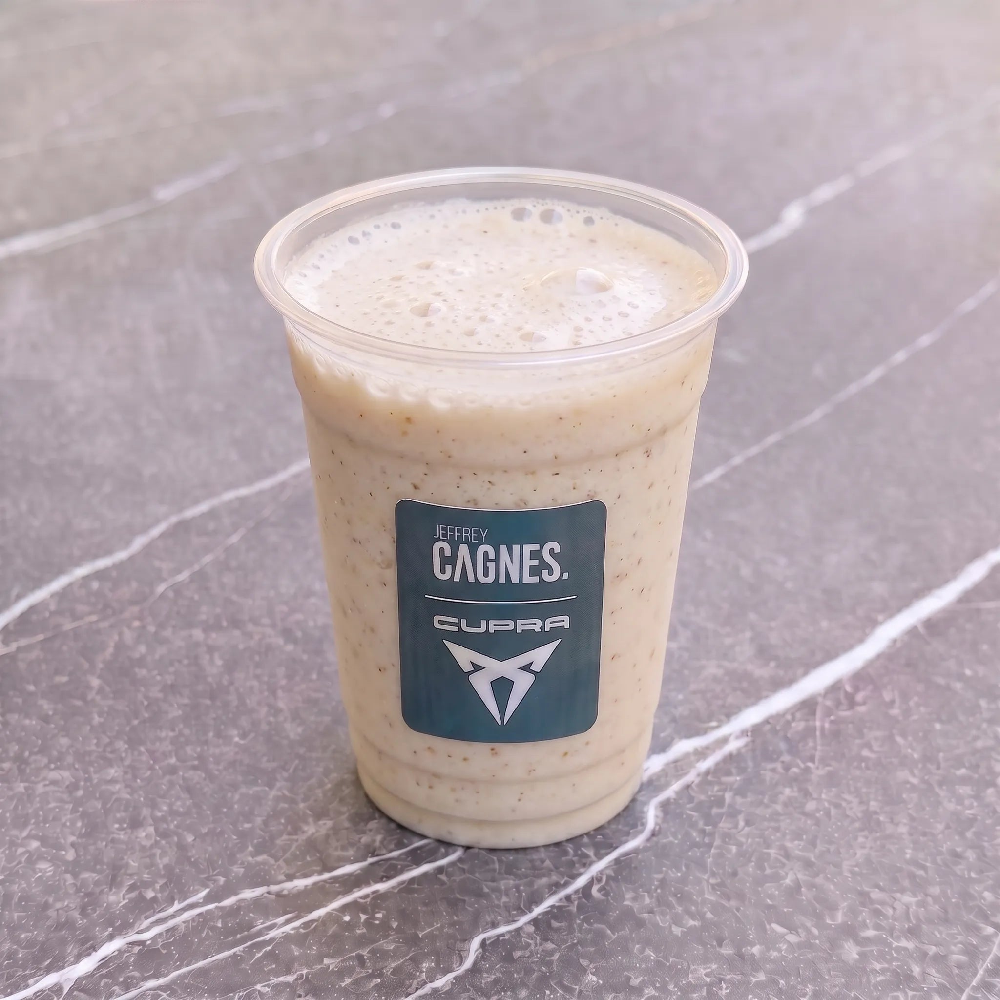 Gobelet transparent de smoothie Cali par Jeffrey Cagnes x Cupra, révélant une texture onctueuse couleur crème parsemée d'éclats de fruits secs et de caramel.
