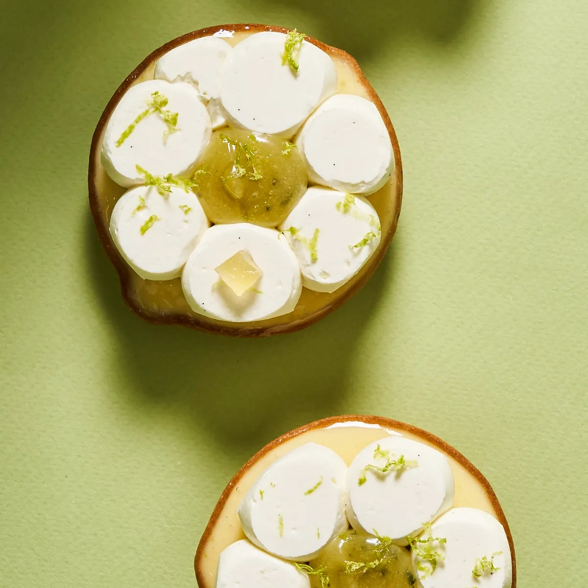 Deux tartes au citron vert individuelles en forme de fleur, ornées de pétales de ganache montée, de zestes frais et d'un cœur de confit vert brillant.