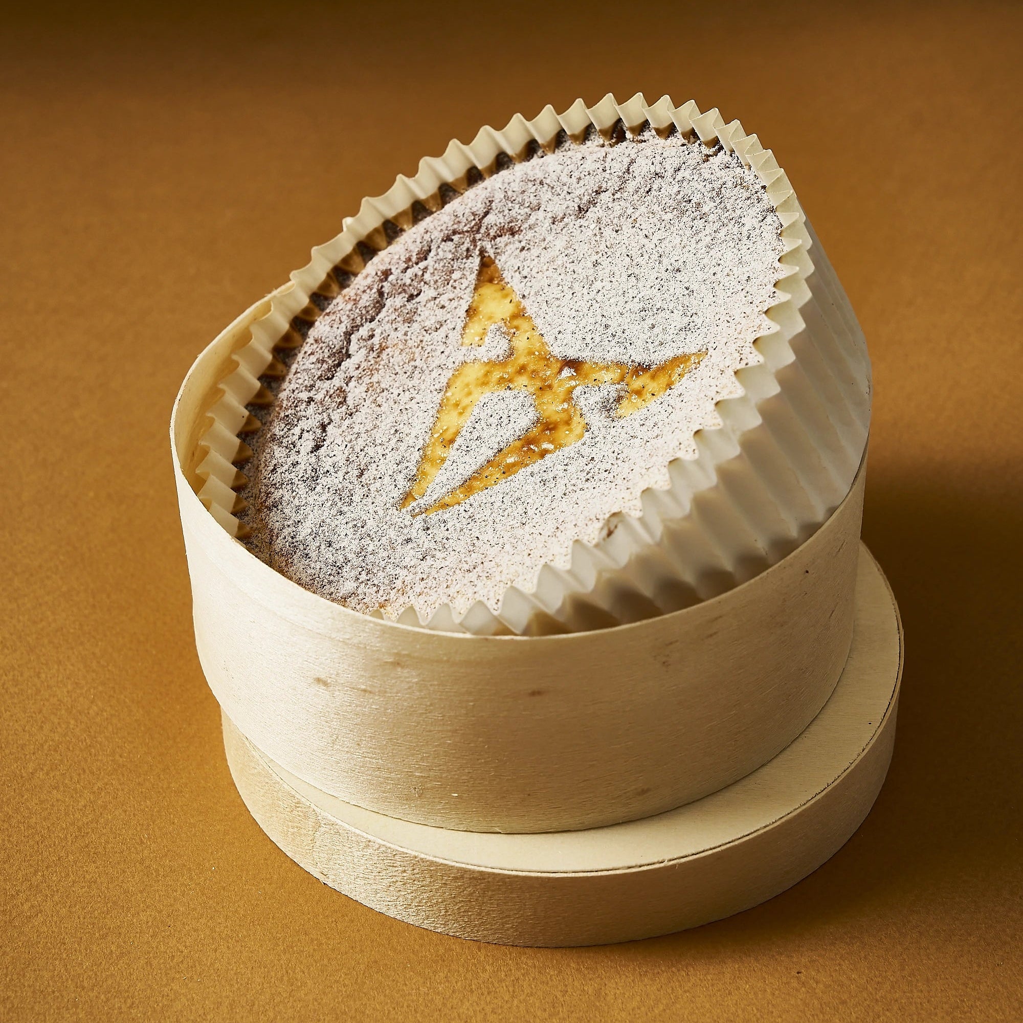 Tarte de Queso exclusive signée Jeffrey Cagnes x Cupra saupoudrée de sucre glace laissant apparaître le logo Cupra, présentée dans sa boîte en bois sur fond ocre.