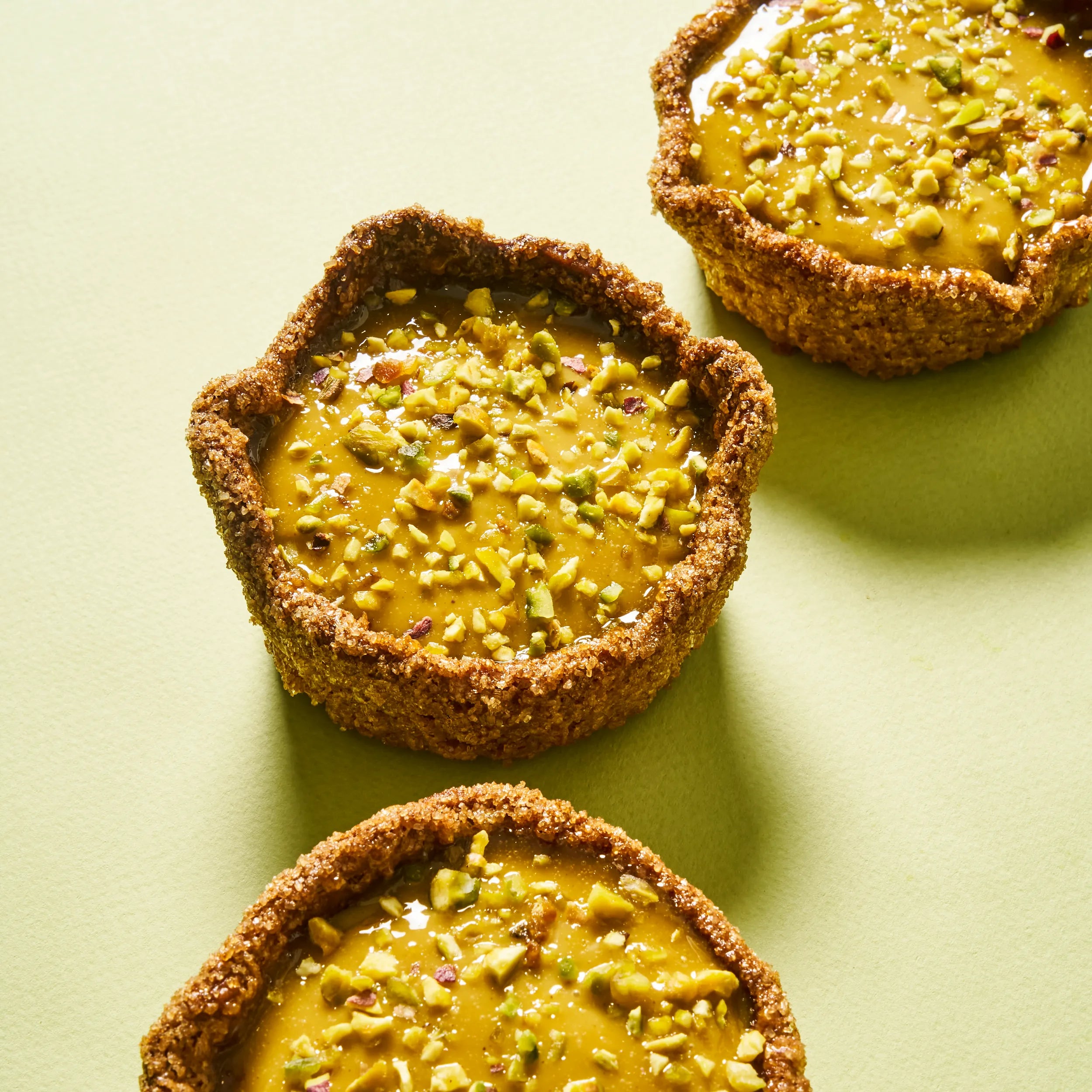 Trois flans à la pistache alignés sur un fond vert amande épuré, illustrant le visuel graphique et gourmand de cette pâtisserie.