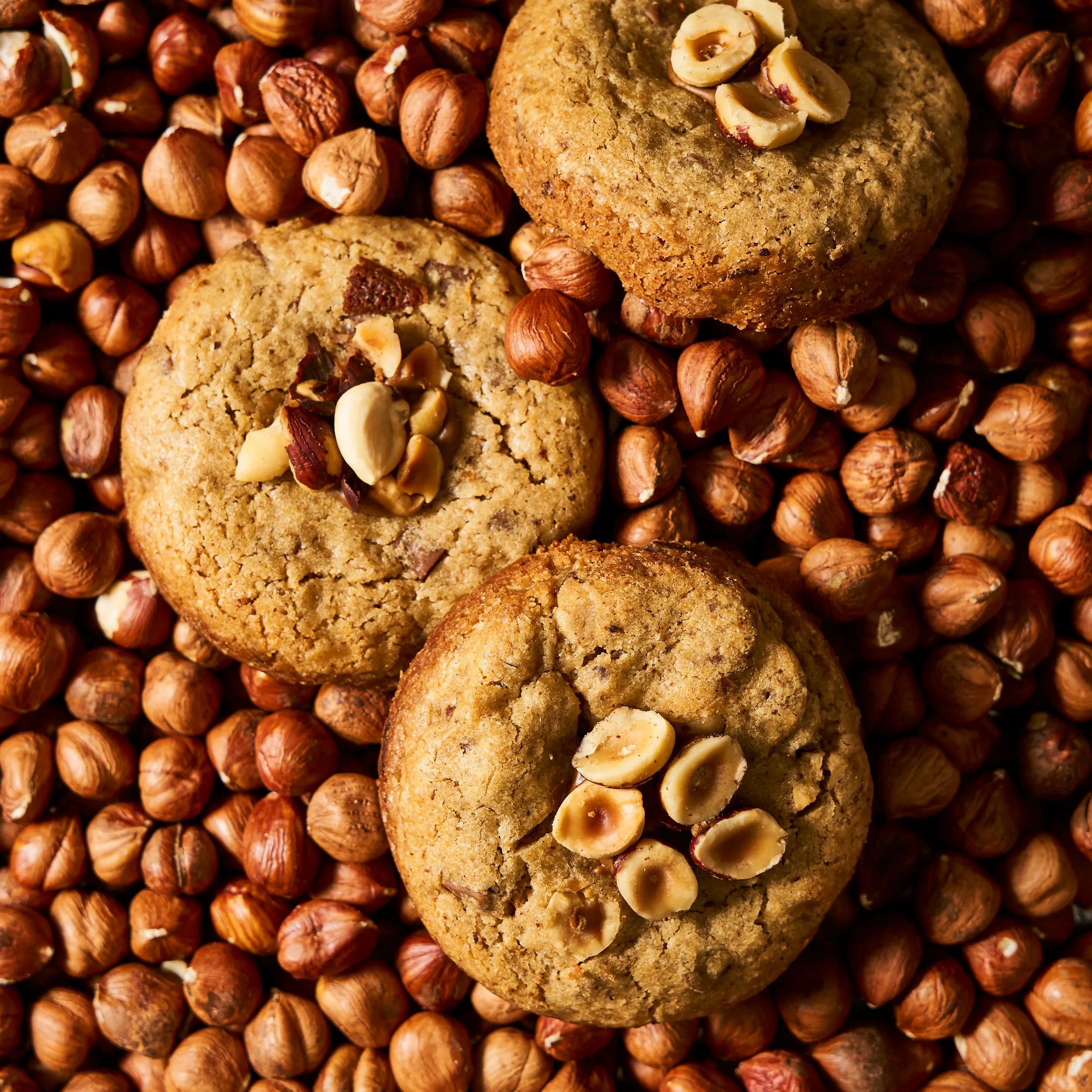 Trois cookies artisanaux chocolat au lait et noisettes torréfiées signés Jeffrey Cagnes, disposés sur un lit de noisettes entières.