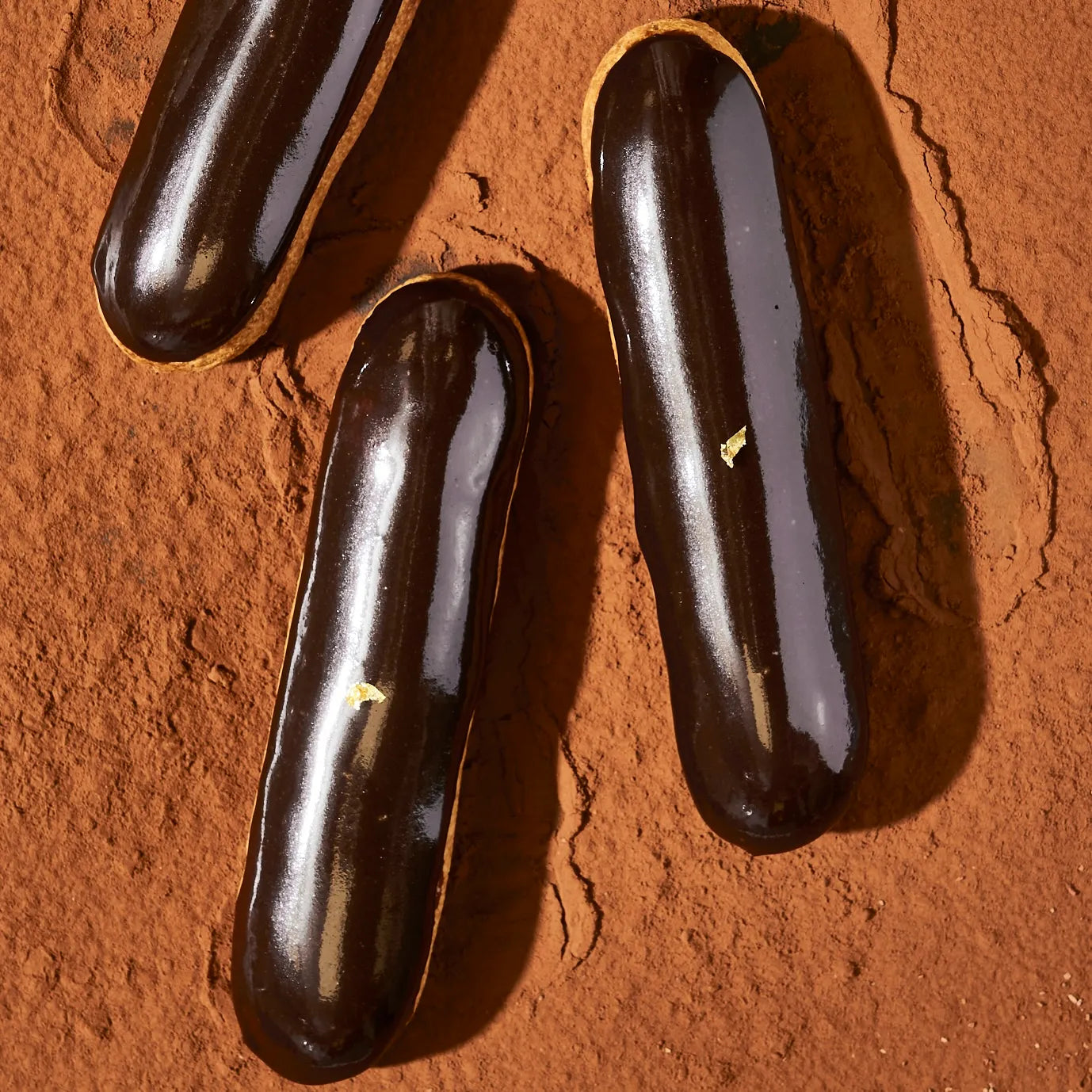 Trois éclairs au chocolat brillants parsemés d'une feuille d'or, reposant sur un fond de poudre de cacao.