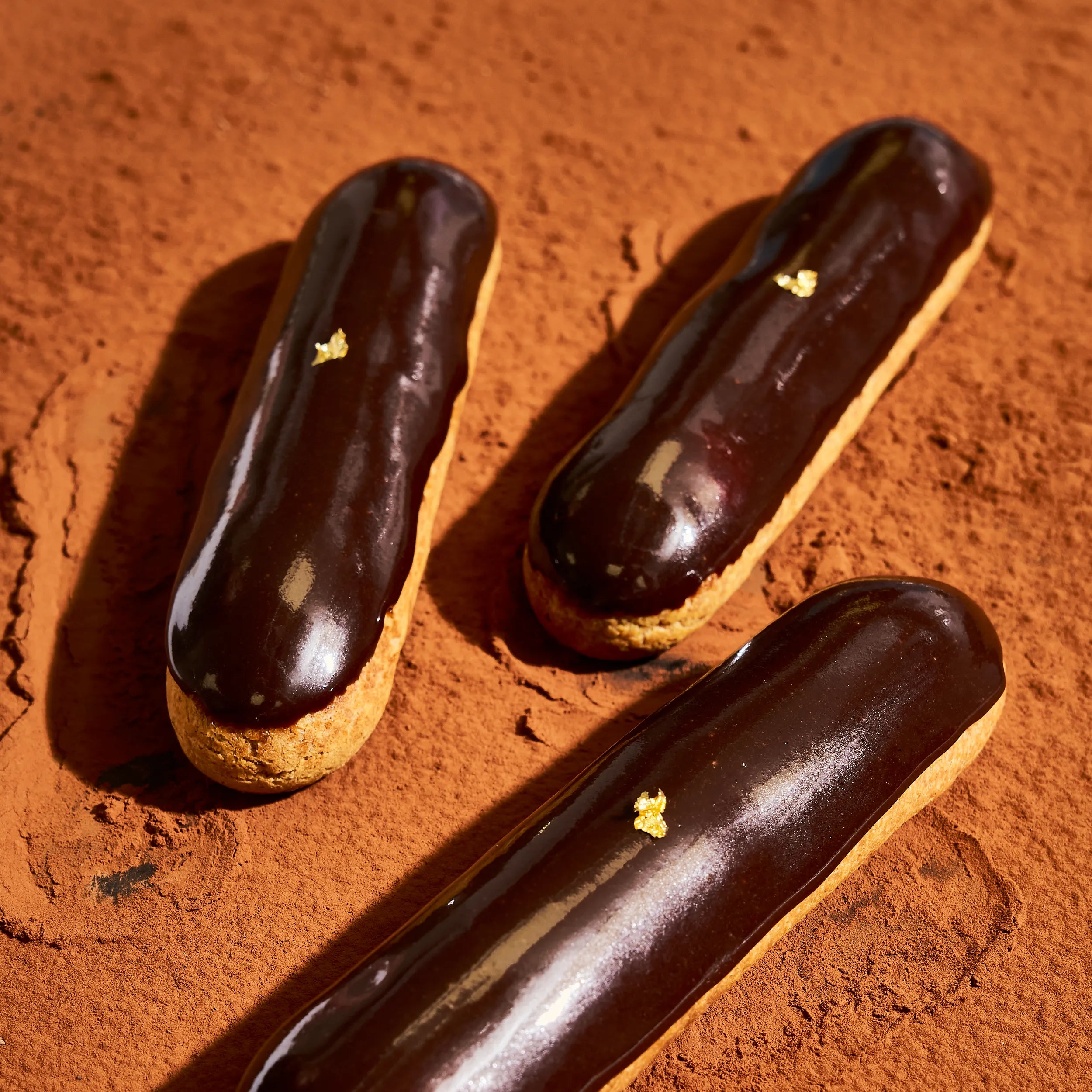 Vue en plongée de trois éclairs au chocolat signés Jeffrey Cagnes sur un lit de cacao ambré.
