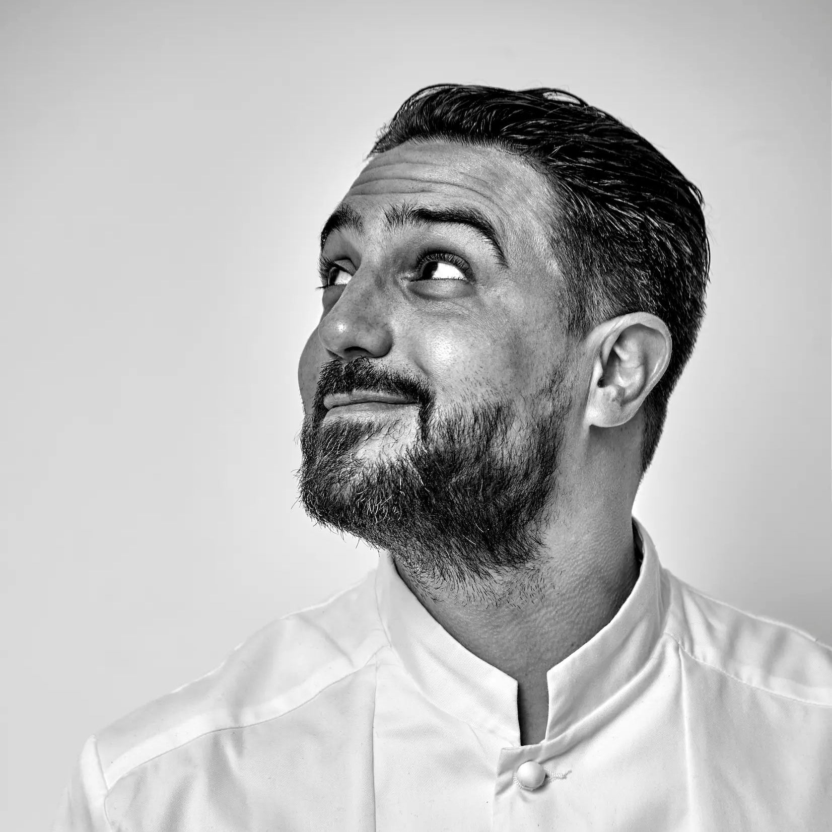 Portrait en buste du Chef Pâtissier Jeffrey Cagnes en veste blanche, regardant vers le haut, sur fond gris clair.