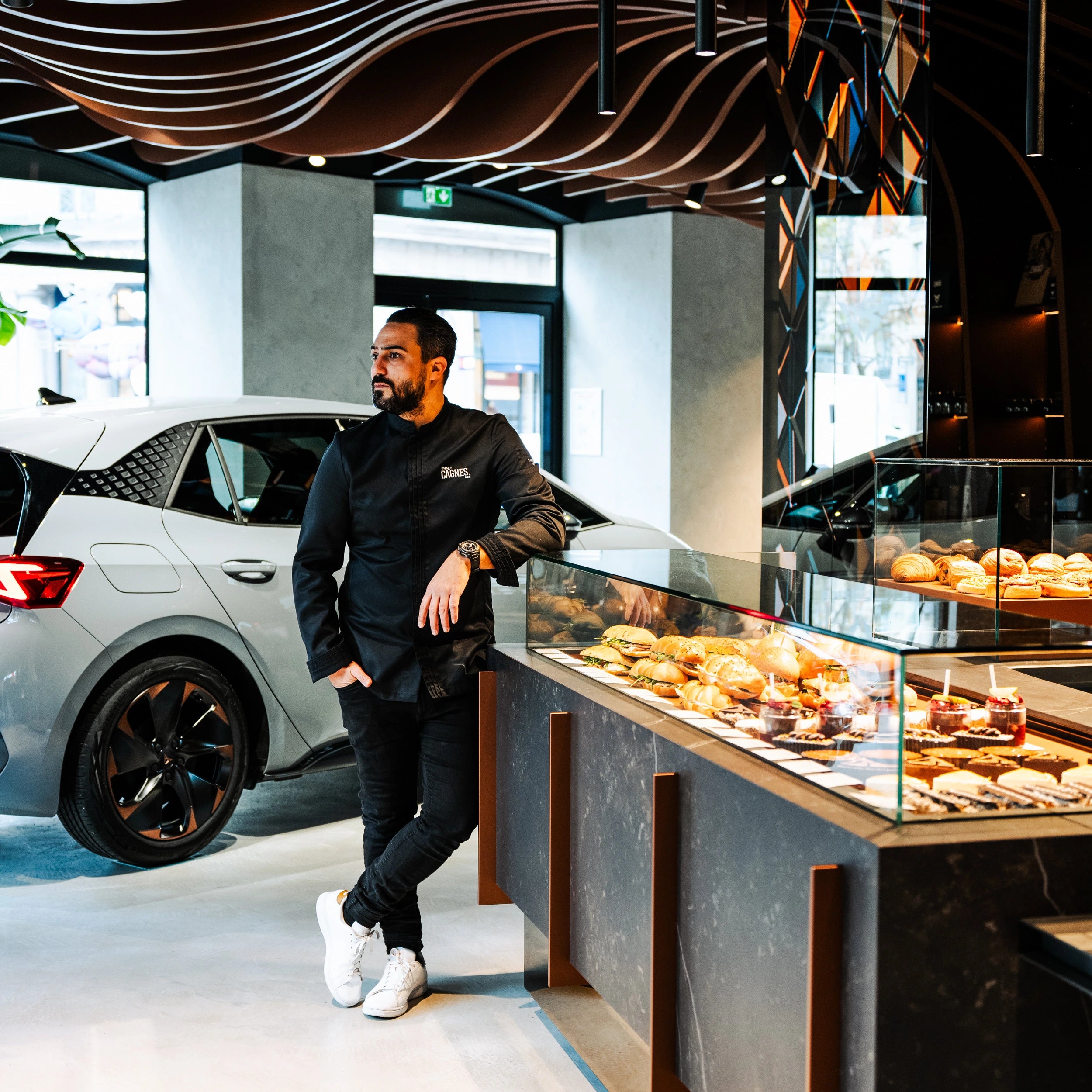 Le Chef Pâtissier Jeffrey Cagnes en veste noire adossé au comptoir vitrine du Coffee Club Cupra au 1 Boulevard de la Madeleine à Paris, à côté d'une voiture Cupra.