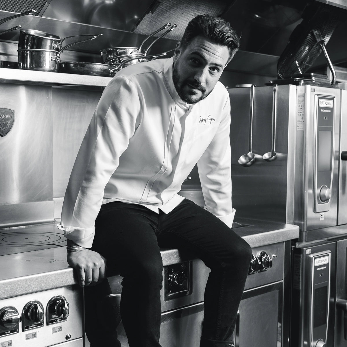 Jeffrey Cagnes assis sur le bord d'un piano de cuisson professionnel, en veste blanche et jean noir, dans sa cuisine.