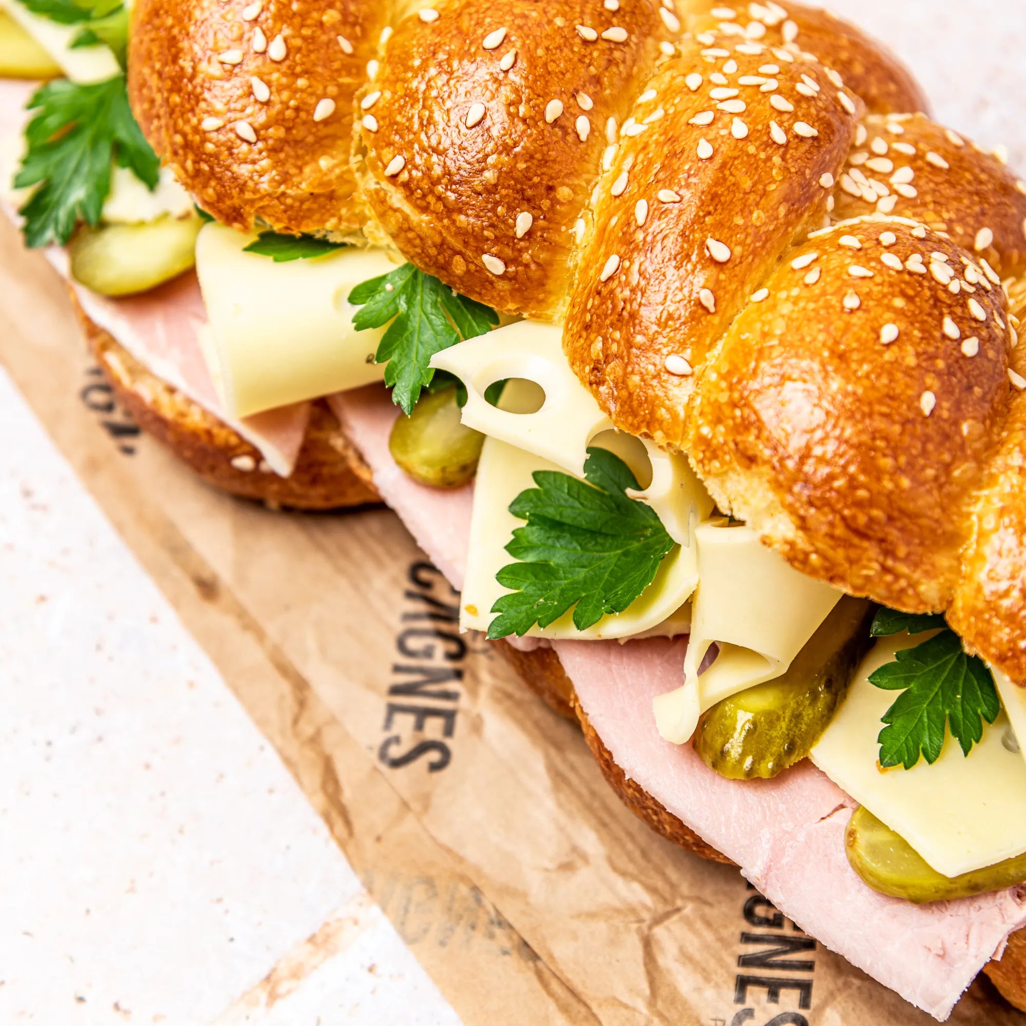 Sandwich Le Parisien tenu en main dans son sachet kraft, révélant la gourmandise du pain Hallot tressé.
