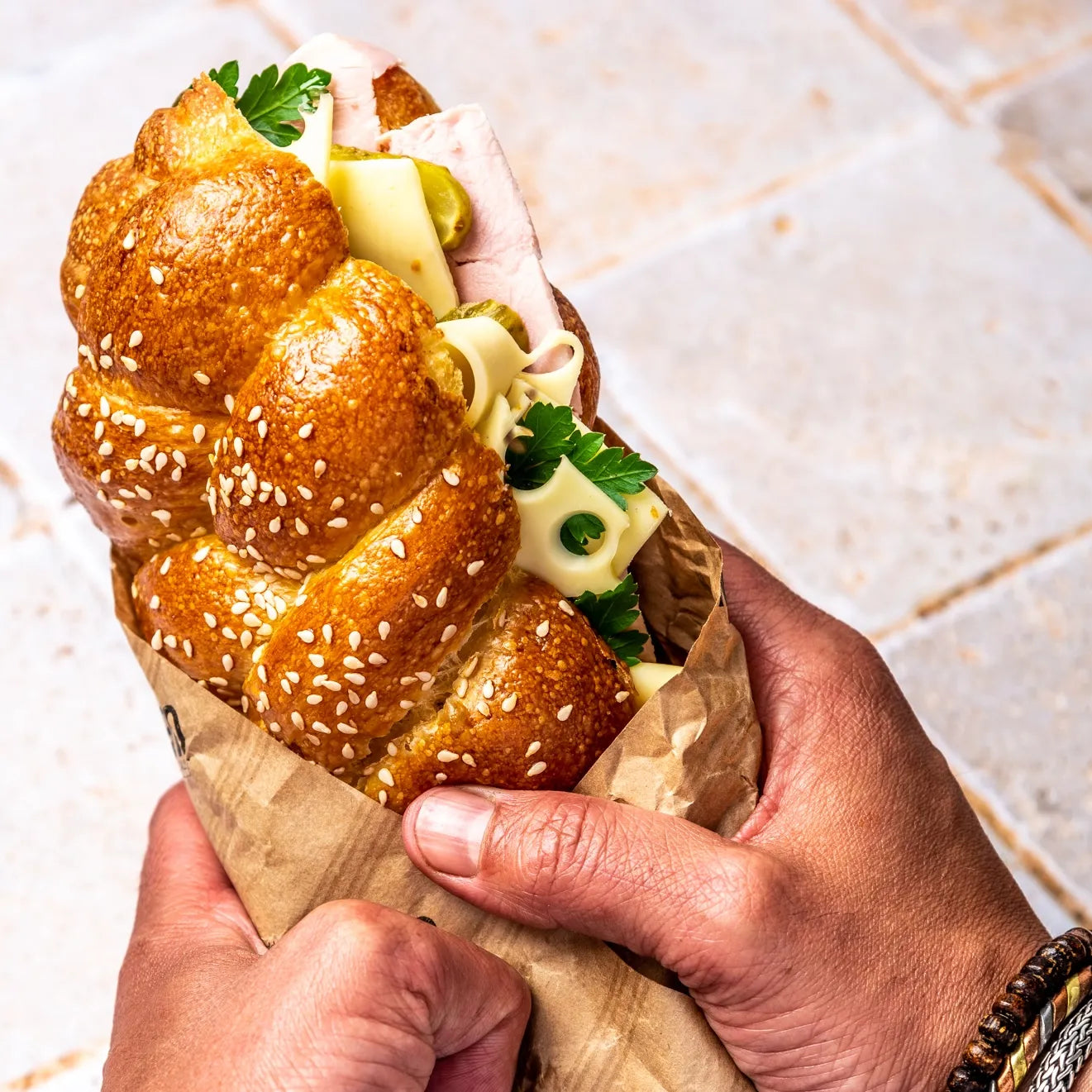 Sandwich Le Parisien tenu en main dans son sachet kraft, révélant la gourmandise du pain Hallot tressé.