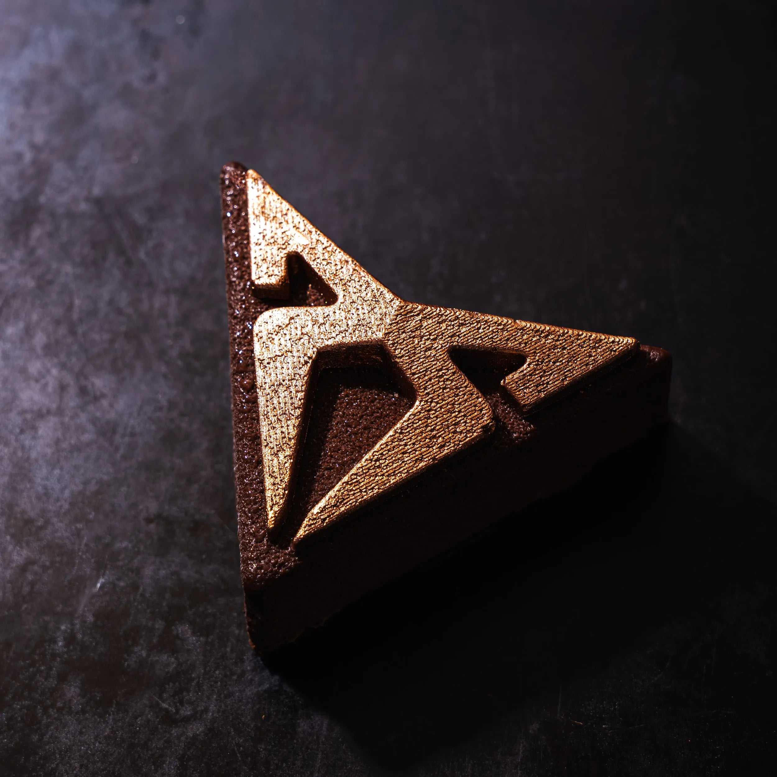 Vue en plongée de l'entremets chocolat au fini mat poudré et logo texturé or, posé sur une plaque de métal patinée.