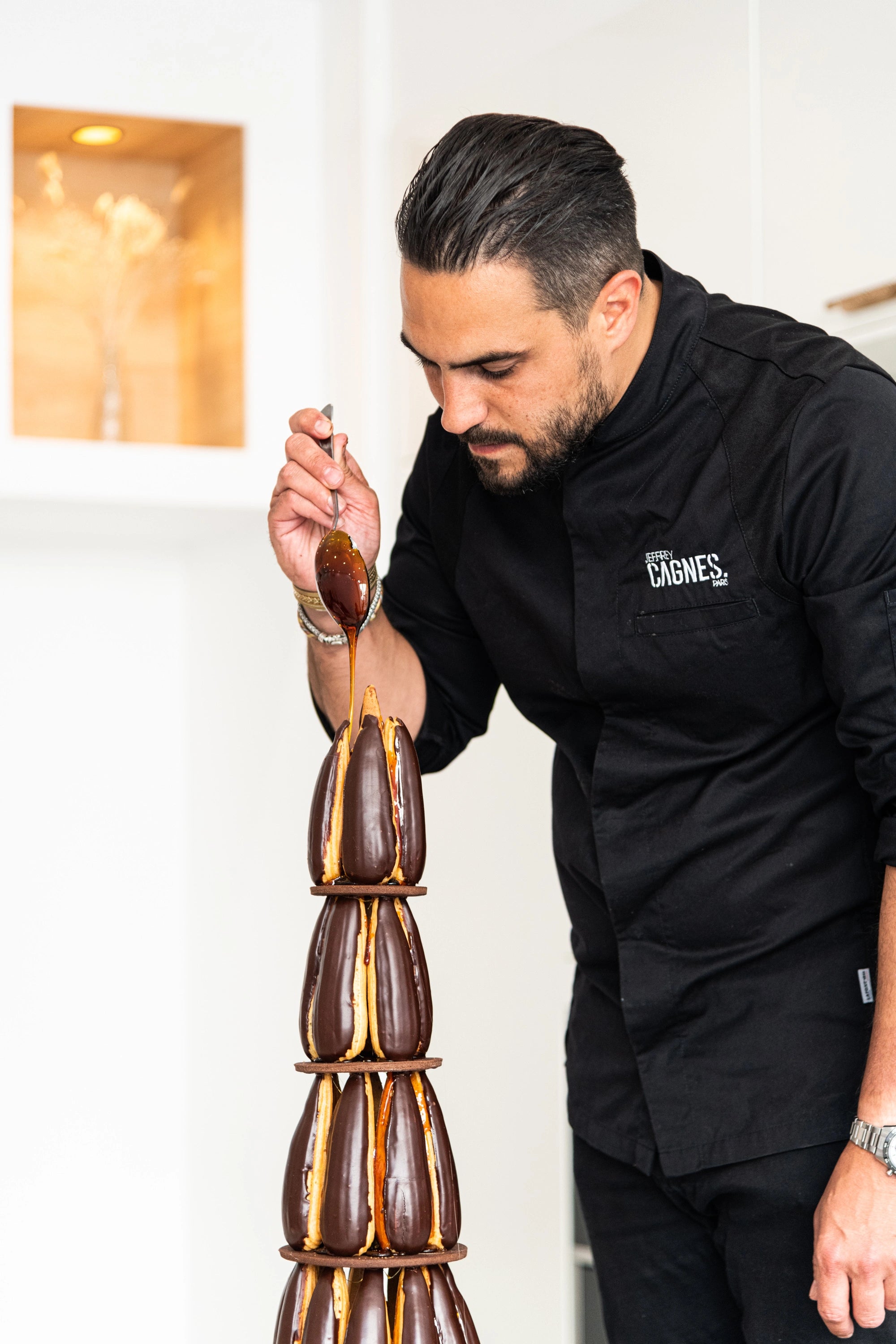 Le Chef pâtissier Jeffrey Cagnes en train d'assembler minutieusement une pièce montée de mariage moderne composée de religieuses et d'éclairs glacés au chocolat avec du caramel filant.