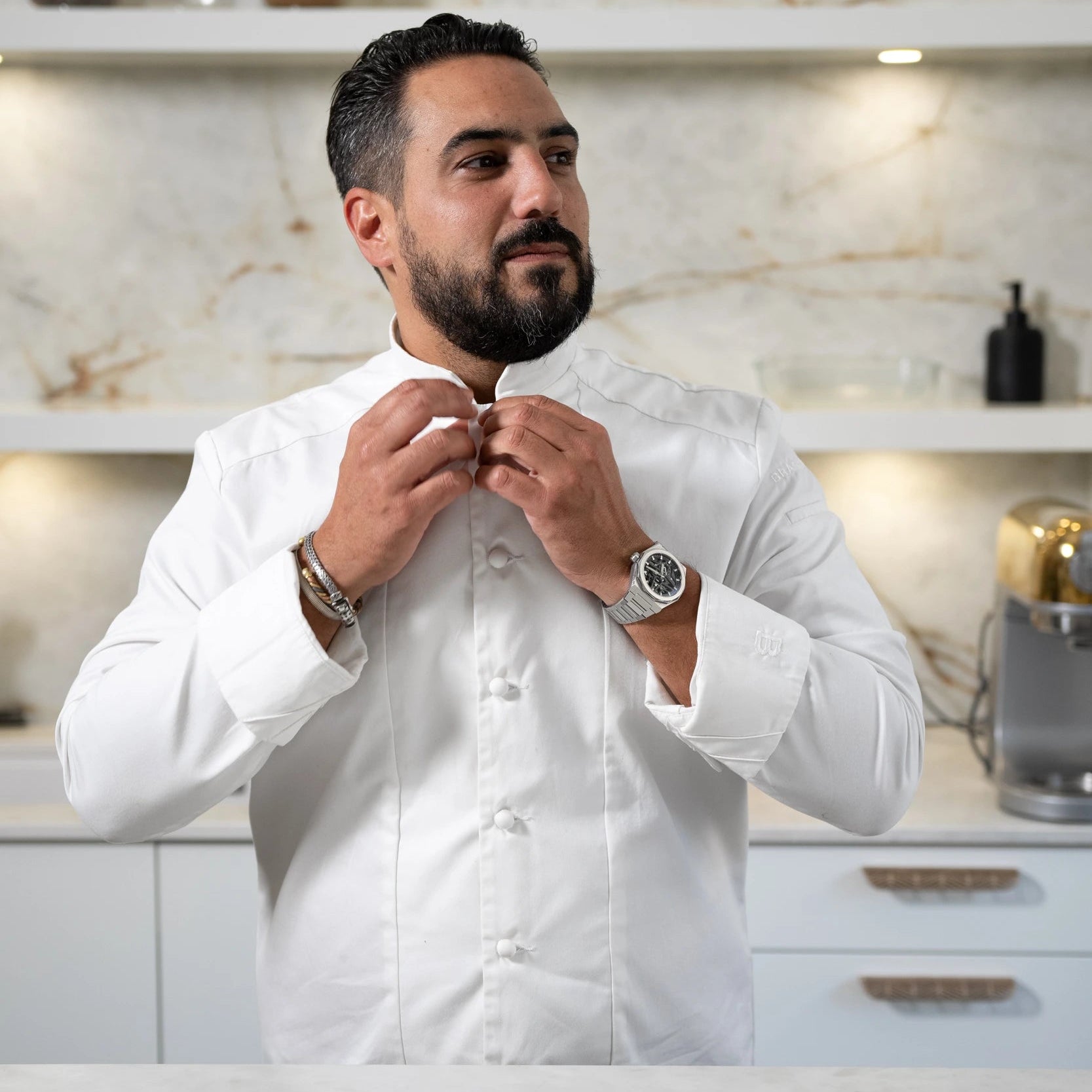 Portrait à la taille du Chef Pâtissier Jeffrey Cagnes ajustant le col de sa veste de cuisine blanche, debout derrière un plan de travail en marbre avec un fouet au premier plan.
