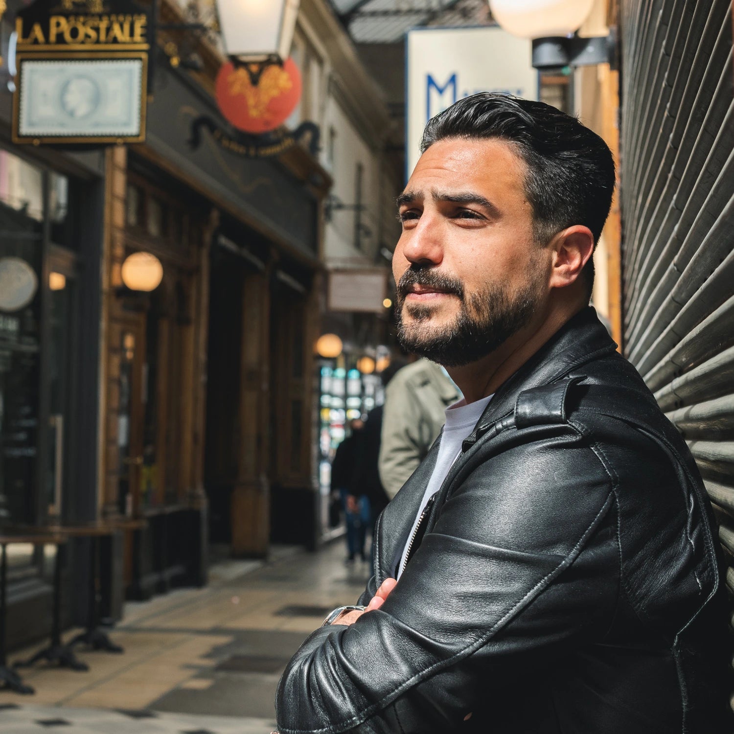 Portrait de Jeffrey Cagnes portant une veste en cuir noire, regardant au loin, debout dans le Passage des Panoramas à Paris, avec des enseignes historiques en arrière-plan.
