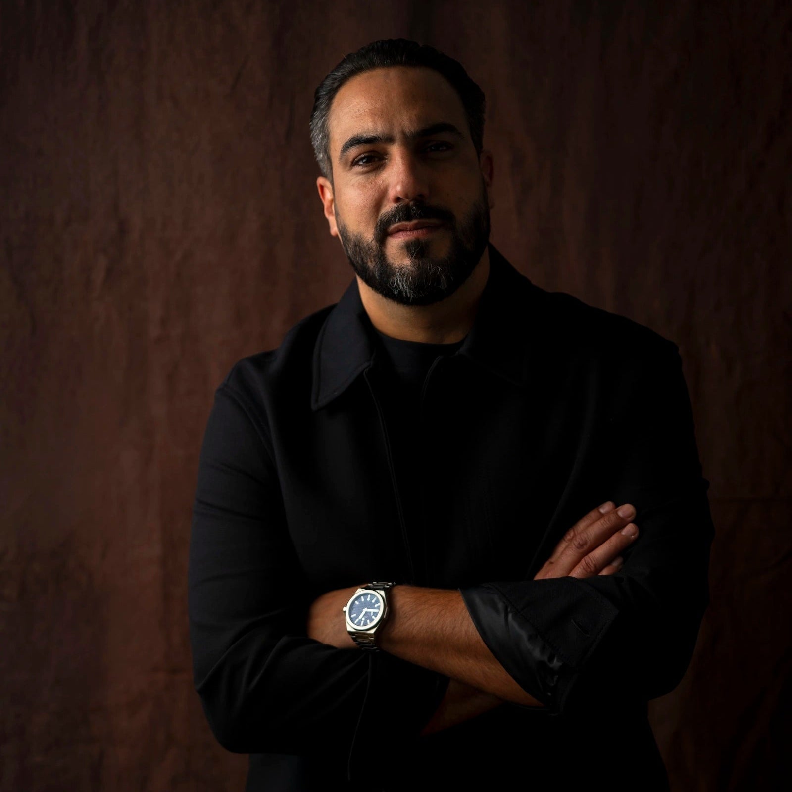 Portrait studio de trois quarts du Chef Jeffrey Cagnes, les bras croisés en veste noire moderne, posant devant un arrière-plan texturé marron foncé avec un éclairage de style clair-obscur.