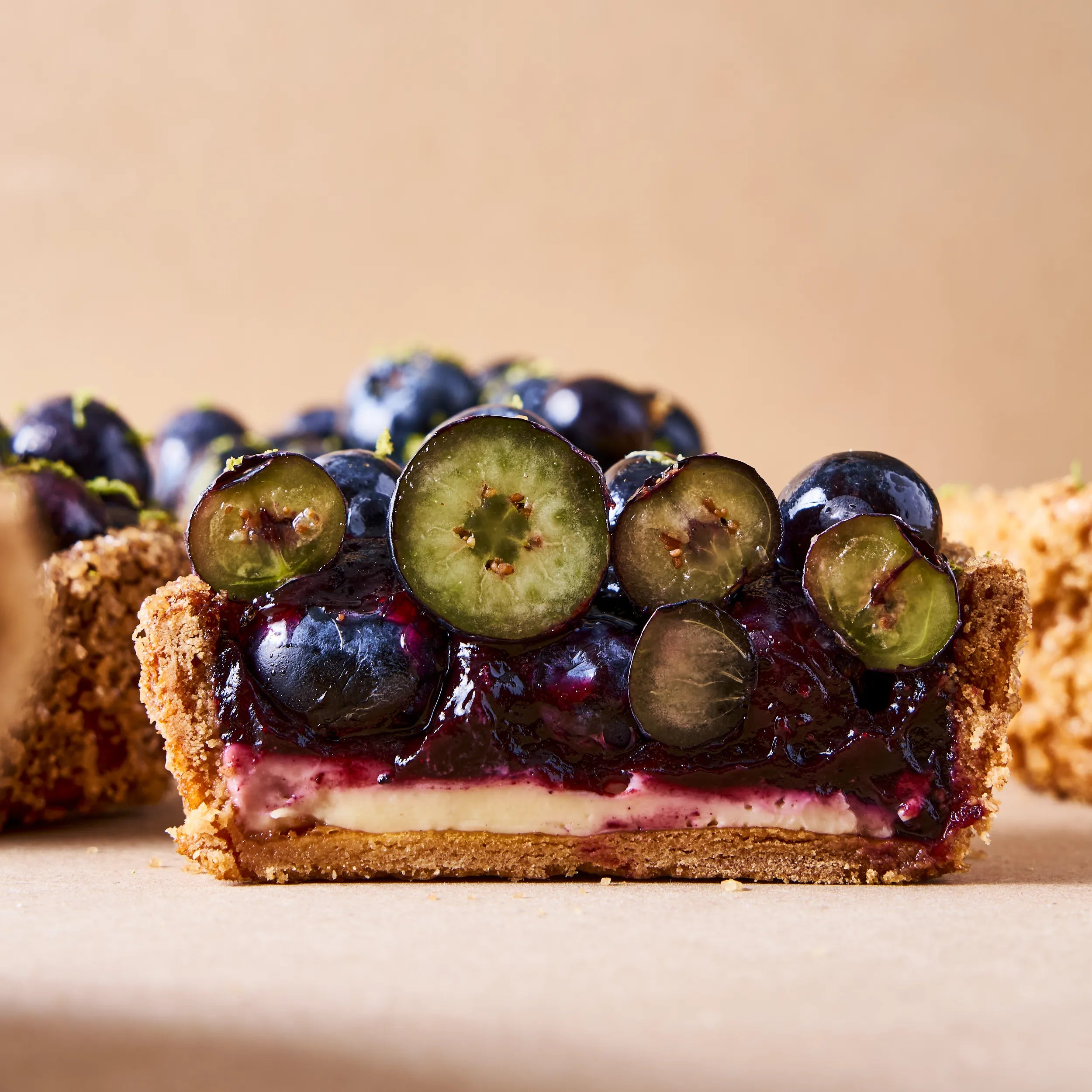 Vue de face de la coupe d'une tartelette aux myrtilles montrant la pâte sucrée dorée, le crémeux vanille et le confit.