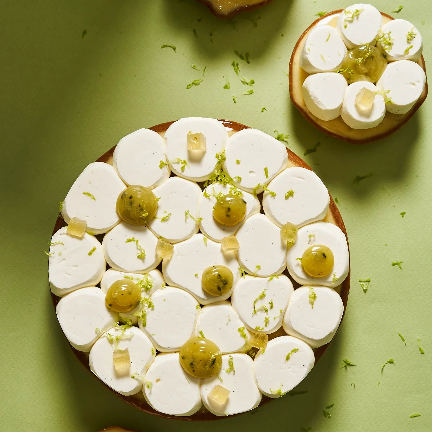 Vue aérienne d'une grande tarte au citron vert de 6 parts et d'une tarte individuelle, parsemées de zestes frais sur un fond vert amande.